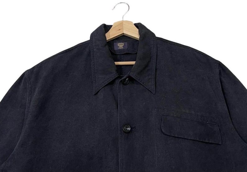 May include: A dark navy blue shirt with a collar and a single pocket, hanging on a wooden hanger. The fabric looks like cotton or denim. The shirt has a classic design. The buttons are black. The shirt is a versatile piece of clothing.