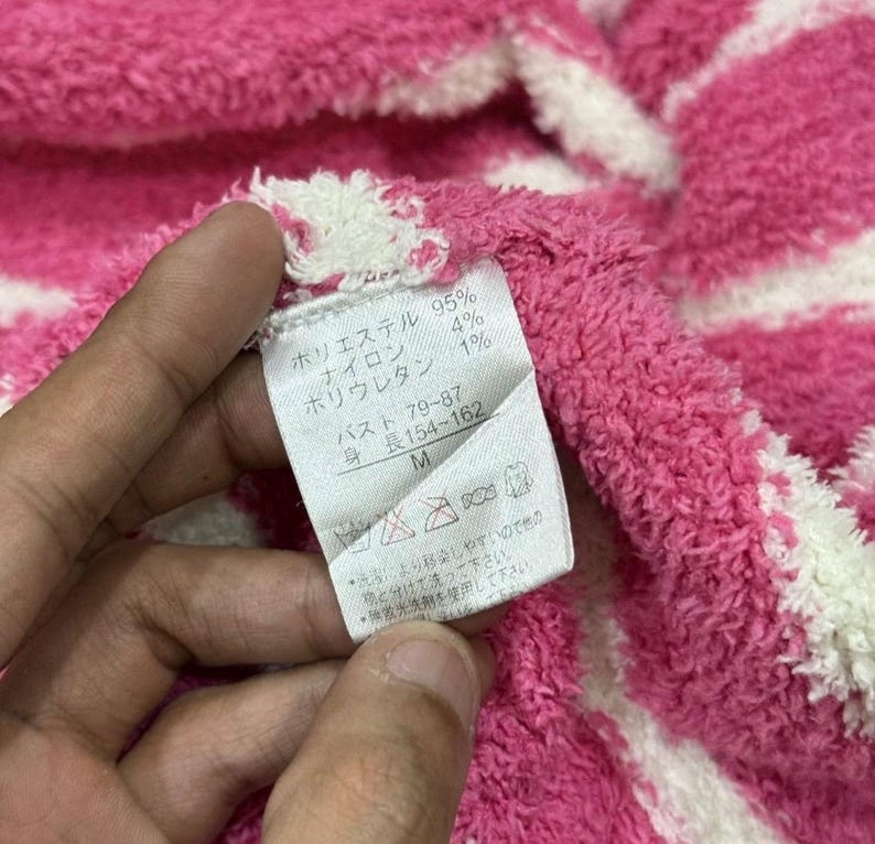 May include: Close-up of a pink and white striped fabric with a tag. The tag has Japanese text indicating the material composition and size. The fabric is soft and textured.