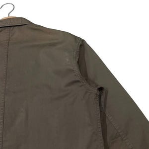 May include: Close-up of a dark olive-green jacket with a collar and long sleeves. The jacket is hanging on a wooden hanger. The fabric appears to be a sturdy cotton or blend, with visible stitching details.