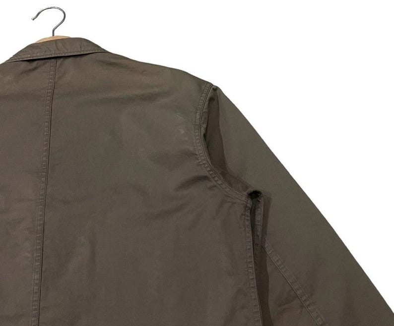 May include: Close-up of a dark olive-green jacket with a collar and long sleeves. The jacket is hanging on a wooden hanger. The fabric appears to be a sturdy cotton or blend, with visible stitching details.