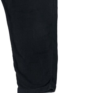 Puede incluir: Primer plano de unos pantalones negros. La tela es de un color oscuro s&oacute;lido, posiblemente con un ligero brillo. Los pantalones est&aacute;n doblados en el dobladillo, creando un borde limpio y acabado. El fondo es blanco.