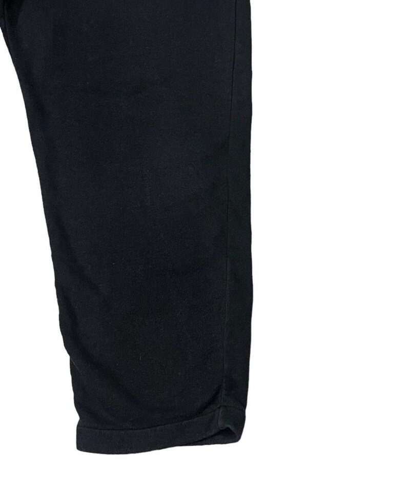 Puede incluir: Primer plano de unos pantalones negros. La tela es de un color oscuro s&oacute;lido, posiblemente con un ligero brillo. Los pantalones est&aacute;n doblados en el dobladillo, creando un borde limpio y acabado. El fondo es blanco.