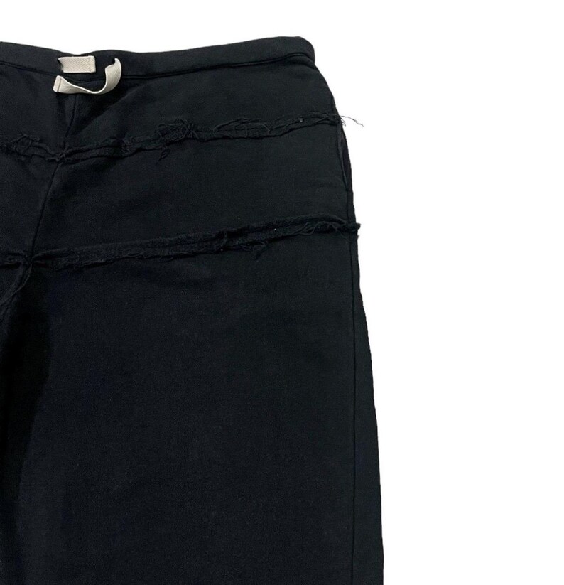 Puede incluir: Pantalones deportivos negros con cintura con cord&oacute;n y detalles desgastados. Los pantalones tienen un corte holgado y presentan costuras horizontales con bordes deshilachados, agregando una textura &uacute;nica. Un cord&oacute;n blanco es visible en la cintura.