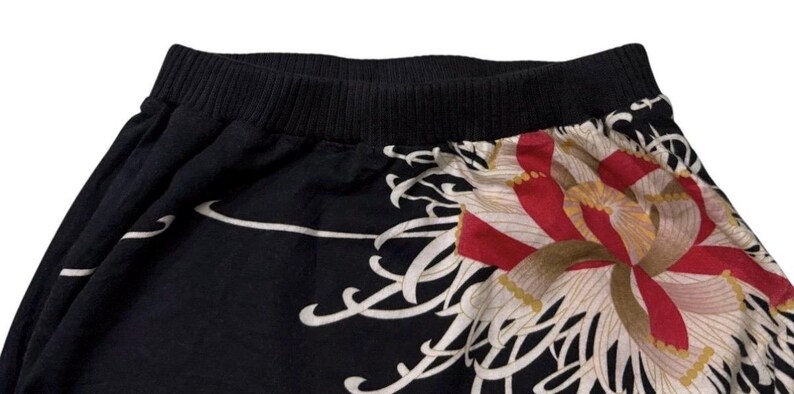 May include: Black skirt with a wide black waistband and a floral design. The skirt features a large, stylized flower in red, white, and gold. White, flowing lines accent the black fabric.