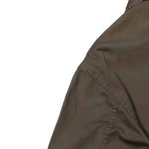 May include: Close-up of a brown shirt, showing the shoulder and part of the sleeve. The fabric has visible stitching along the seams. The shirt is hanging on a wooden hanger.