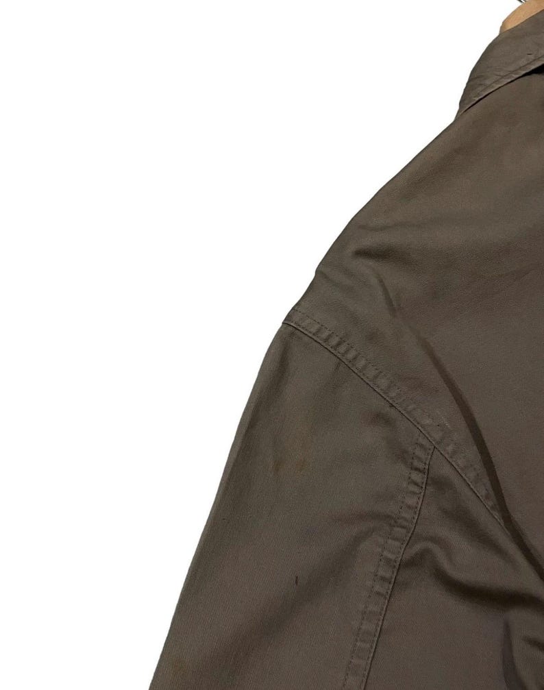 May include: Close-up of a brown shirt, showing the shoulder and part of the sleeve. The fabric has visible stitching along the seams. The shirt is hanging on a wooden hanger.