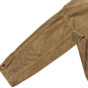 K&ouml;nnte beinhalten: Nahaufnahme eines beigefarbenen Cordjacken&auml;rmels. Der &Auml;rmel hat eine gekn&ouml;pfte Manschette und sichtbare N&auml;hte entlang der N&auml;hte. Der Cordstoff hat eine gerippte Textur.