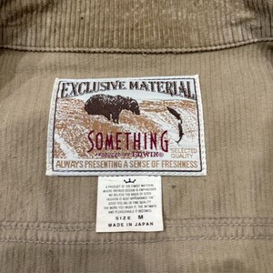 K&ouml;nnte beinhalten: Nahaufnahme einer beigefarbenen Cordjacke mit einem Etikett mit der Aufschrift "EXCLUSIVE MATERIAL SOMETHING". Das Etikett enth&auml;lt auch den Text "ALWAYS PRESENTING A SENSE OF FRESHNESS". Die Jacke ist Gr&ouml;&szlig;e M und hergestellt in Japan.
