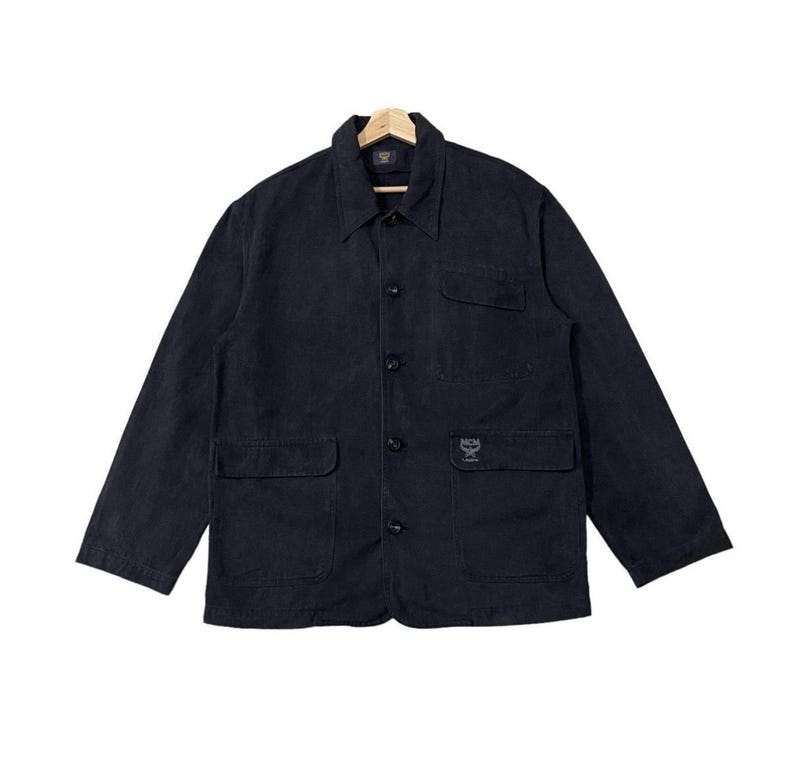 May include: A dark navy blue jacket with a collared neckline, button-down front, and two front pockets. The jacket has long sleeves and a small logo on the front pocket. The jacket is hanging on a wooden hanger.