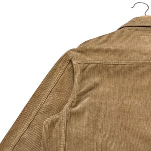 K&ouml;nnte beinhalten: Nahaufnahme einer hellbraunen Cordjacke, die an einem Holzb&uuml;gel h&auml;ngt. Die Jacke hat ein Knopfdesign, einen Kragen und vertikale Cordrippen. Der Stoff hat ein weiches, strukturiertes Aussehen.