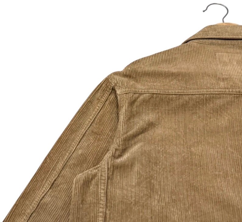 K&ouml;nnte beinhalten: Nahaufnahme einer hellbraunen Cordjacke, die an einem Holzb&uuml;gel h&auml;ngt. Die Jacke hat ein Knopfdesign, einen Kragen und vertikale Cordrippen. Der Stoff hat ein weiches, strukturiertes Aussehen.