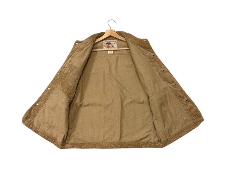 K&ouml;nnte beinhalten: Offene, hellbraune Cordjacke mit Knopfverschluss und zwei Vordertaschen. Sie h&auml;ngt an einem Holzkleiderb&uuml;gel. Das Innenfutter ist heller. Auf der Innenseite befindet sich ein Etikett.