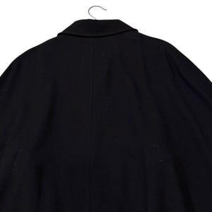 May include: A black coat with a notched collar, hanging from a metal hanger. The coat is made of a dark fabric and has a simple, classic design. The back of the coat is visible, showing a seam down the center.