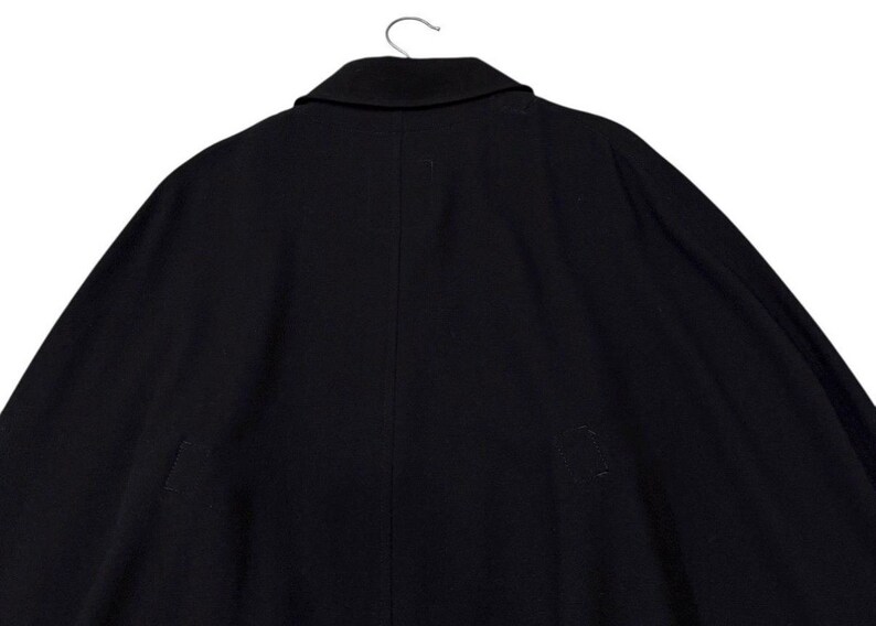 May include: A black coat with a notched collar, hanging from a metal hanger. The coat is made of a dark fabric and has a simple, classic design. The back of the coat is visible, showing a seam down the center.
