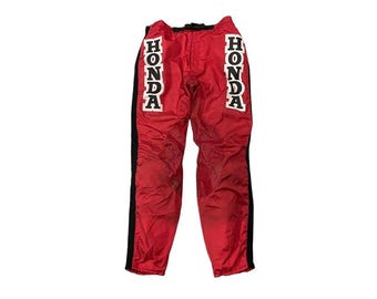 May include: Red racing pants with the word "HONDA" printed vertically in white on both legs. The pants have black stripes down the sides and a black waistband. These pants are likely for motorcycle racing or other motorsports.