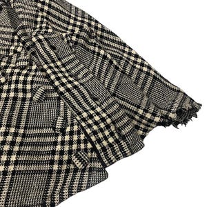 May include: Close-up of a black and white patterned blazer. The fabric features a classic houndstooth design with a frayed edge on the sleeve. The blazer is made of a woven material.