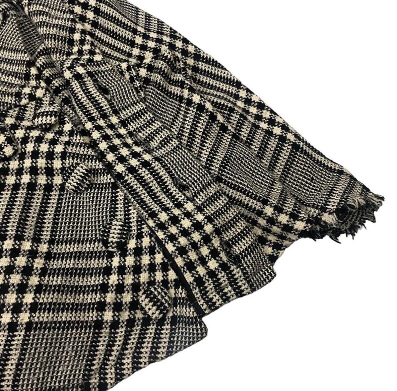 May include: Close-up of a black and white patterned blazer. The fabric features a classic houndstooth design with a frayed edge on the sleeve. The blazer is made of a woven material.