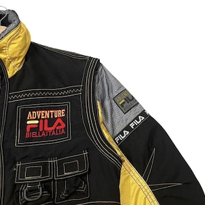 以下が含まれることがあります： 前面に「Adventure Fila Biellaitlia」の文字が入った、黒と黄色のFila Adventureジャケット。 ジャケットには2つのフロントポケットと黄色のステッチが施されています。 ジャケットにはグレーと黒のショルダーパネルが付いています。