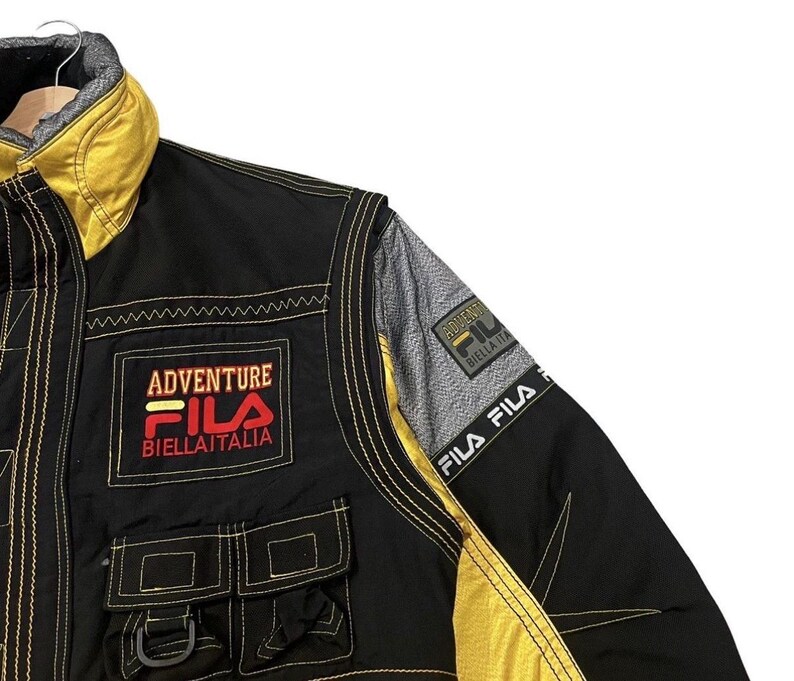 以下が含まれることがあります： 前面に「Adventure Fila Biellaitlia」の文字が入った、黒と黄色のFila Adventureジャケット。 ジャケットには2つのフロントポケットと黄色のステッチが施されています。 ジャケットにはグレーと黒のショルダーパネルが付いています。