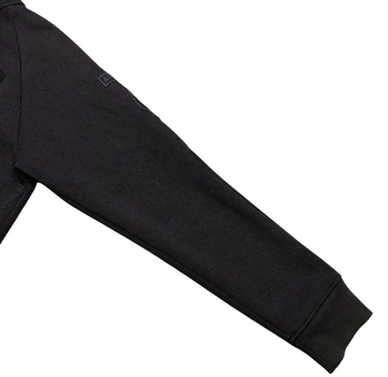 May include: Close-up of a dark grey sweatshirt sleeve. The sleeve has a ribbed cuff and a small square patch near the shoulder. The fabric appears to be a textured knit.