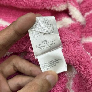 May include: Close-up of a pink and white striped fabric with a tag held by a hand. The tag has Japanese text, a phone number, and the Universal Studios Japan logo. The fabric appears soft and textured.