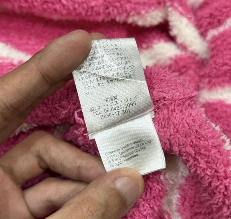 May include: Close-up of a pink and white striped fabric with a tag held by a hand. The tag has Japanese text, a phone number, and the Universal Studios Japan logo. The fabric appears soft and textured.