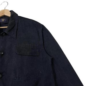 May include: A dark navy blue jacket with a button-down front, a collared neckline, and a chest pocket. The jacket is hanging on a wooden hanger. The MCM logo is visible on the label.