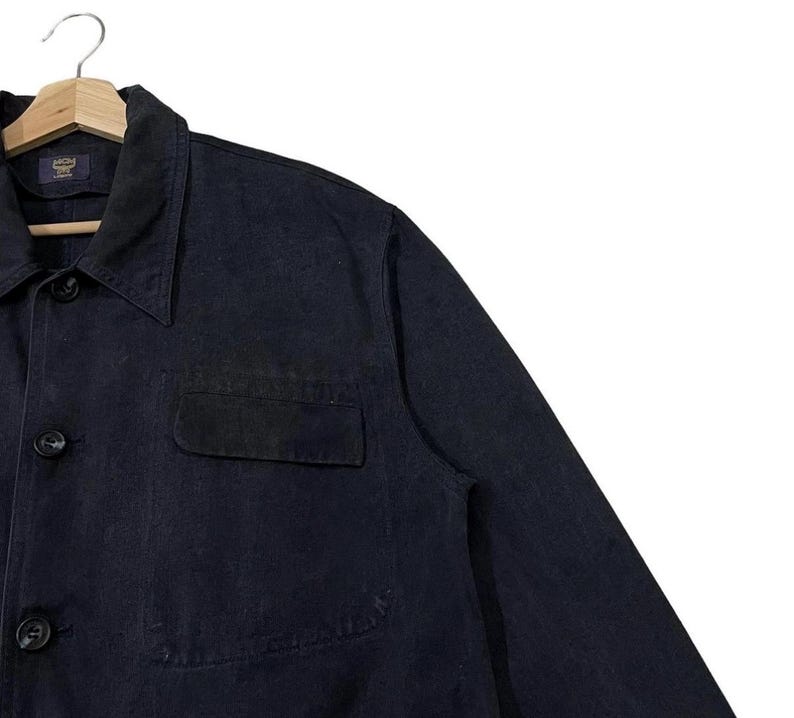 May include: A dark navy blue jacket with a button-down front, a collared neckline, and a chest pocket. The jacket is hanging on a wooden hanger. The MCM logo is visible on the label.