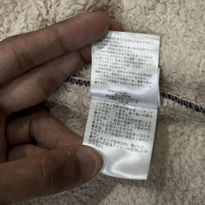 May include: Close-up of a cream-coloured, textured fabric with a black trim. A white tag with Japanese text is attached to the fabric. The tag contains text and care instructions.
