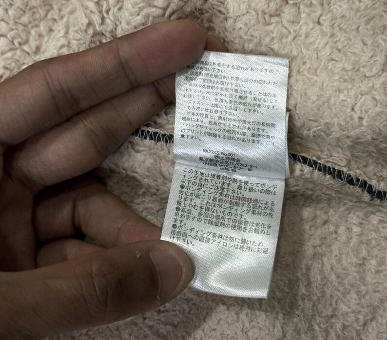 May include: Close-up of a cream-coloured, textured fabric with a black trim. A white tag with Japanese text is attached to the fabric. The tag contains text and care instructions.