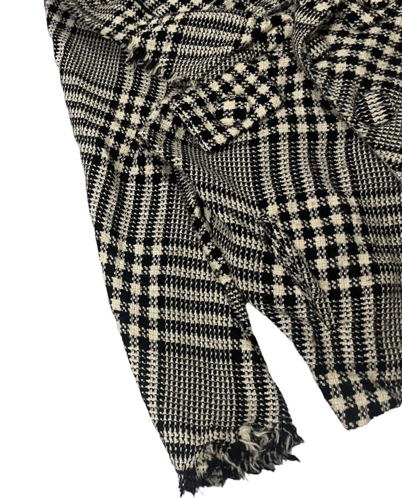 May include: Close-up of a black and white houndstooth patterned fabric. The fabric has a classic check design with a frayed edge. The material appears to be woven, with a textured surface. The overall look is a vintage or retro style.