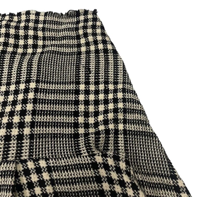 May include: Close-up of a black and white plaid skirt. The skirt has a classic pattern with a repeating grid design. The fabric appears to be a woven material. The skirt's hemline is visible, and the overall style is timeless.