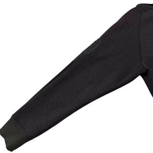 May include: Close-up of a dark grey sweatshirt sleeve. The sleeve features a ribbed cuff and a small, dark patch on the upper arm. The fabric appears to be a soft, textured material.