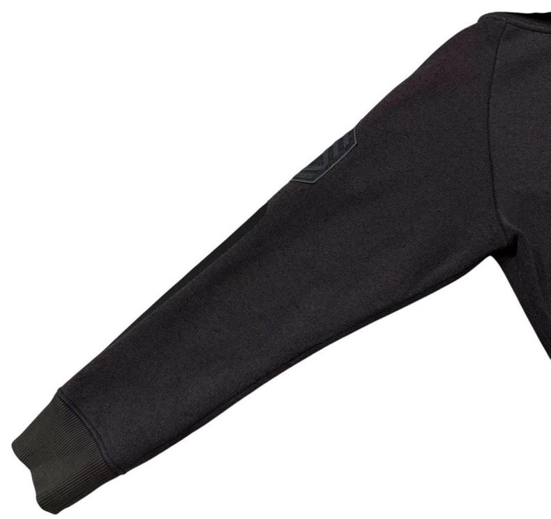 May include: Close-up of a dark grey sweatshirt sleeve. The sleeve features a ribbed cuff and a small, dark patch on the upper arm. The fabric appears to be a soft, textured material.