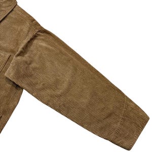 K&ouml;nnte beinhalten: Nahaufnahme eines tan-farbenen Cordjacken&auml;rmels. Der Stoff hat eine gerippte Textur und einen warmen, erdigen Ton. Die Jacke scheint aus einem dicken, strapazierf&auml;higen Material gefertigt zu sein, was darauf hindeutet, dass sie f&uuml;r k&auml;lteres Wetter konzipiert ist.