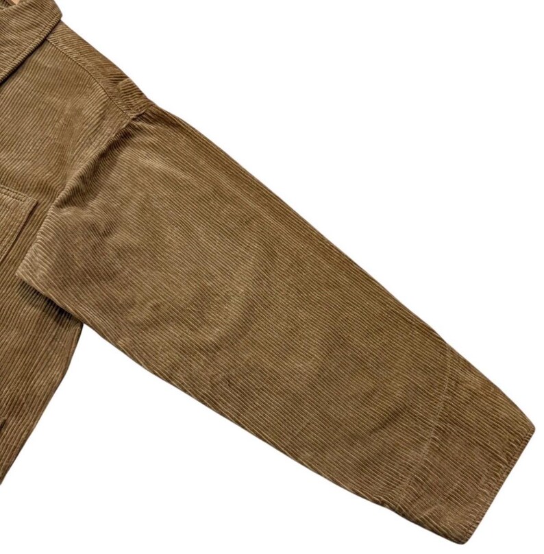 K&ouml;nnte beinhalten: Nahaufnahme eines tan-farbenen Cordjacken&auml;rmels. Der Stoff hat eine gerippte Textur und einen warmen, erdigen Ton. Die Jacke scheint aus einem dicken, strapazierf&auml;higen Material gefertigt zu sein, was darauf hindeutet, dass sie f&uuml;r k&auml;lteres Wetter konzipiert ist.