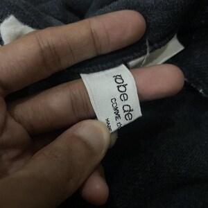 Puede incluir: Primer plano de una etiqueta blanca con el texto "robe de comme de" en negro, con la palabra "MADE" debajo. La etiqueta est&aacute; adherida a una prenda gris oscuro.