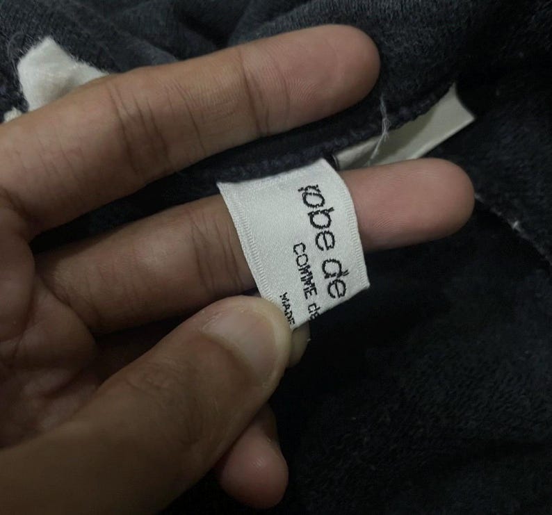 Puede incluir: Primer plano de una etiqueta blanca con el texto "robe de comme de" en negro, con la palabra "MADE" debajo. La etiqueta est&aacute; adherida a una prenda gris oscuro.