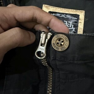 Puede incluir: Primer plano de una cremallera de chaqueta negra con un tirador de cremallera de metal dorado. Se ve un bot&oacute;n dorado con las palabras "Von Dutch". La chaqueta tiene una etiqueta con el logotipo de Von Dutch.