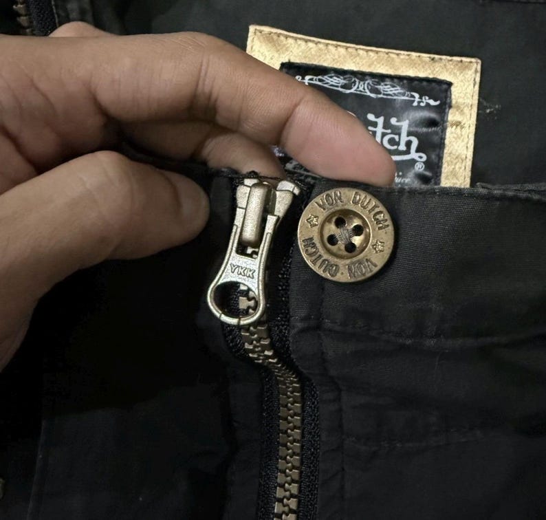 Puede incluir: Primer plano de una cremallera de chaqueta negra con un tirador de cremallera de metal dorado. Se ve un bot&oacute;n dorado con las palabras "Von Dutch". La chaqueta tiene una etiqueta con el logotipo de Von Dutch.