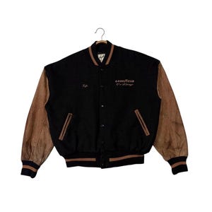 May include: Black and brown varsity jacket with leather sleeves. The jacket features the text "GOOD YEAR" and "Kyle" embroidered on the front. The jacket has a button-up closure and striped cuffs and hem.