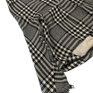 May include: Black and white plaid fabric with a fringed edge. The fabric has a classic houndstooth pattern, with a mix of black and off-white squares and lines. The fabric is folded, revealing a glimpse of a beige lining.