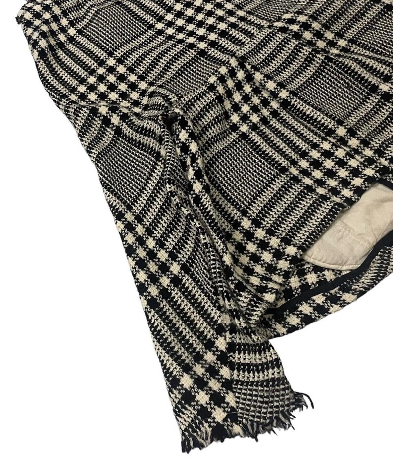May include: Black and white plaid fabric with a fringed edge. The fabric has a classic houndstooth pattern, with a mix of black and off-white squares and lines. The fabric is folded, revealing a glimpse of a beige lining.