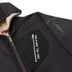 May include: Dark grey hooded zip-up jacket with a black pocket and the text "FREE FALL JUMP IN MIND." The jacket features a black patch on the sleeve with the word "AIRBORNE" and a military emblem. The interior lining of the hood is a light beige.