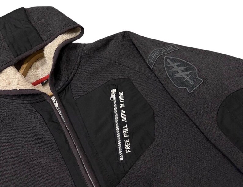 May include: Dark grey hooded zip-up jacket with a black pocket and the text "FREE FALL JUMP IN MIND." The jacket features a black patch on the sleeve with the word "AIRBORNE" and a military emblem. The interior lining of the hood is a light beige.
