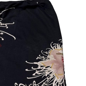 May include: Black fabric with a floral design. The design features a large, stylized flower in shades of pink, beige, and red, with white, flowing accents. The fabric appears to be a skirt or trousers.