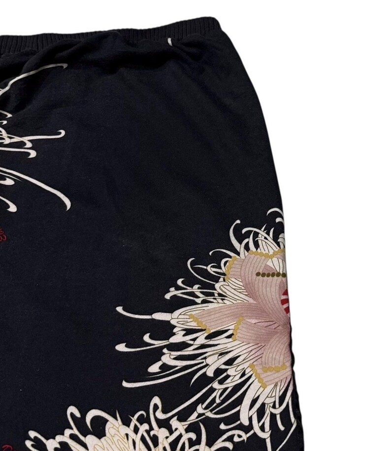 May include: Black fabric with a floral design. The design features a large, stylized flower in shades of pink, beige, and red, with white, flowing accents. The fabric appears to be a skirt or trousers.