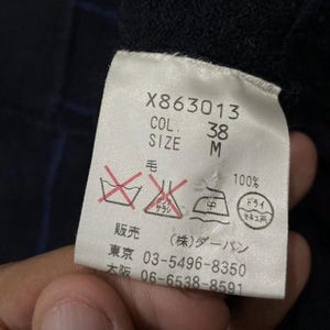 Puede incluir: Primer plano de una etiqueta de ropa con el texto "X863013", "COL. 38" y "SIZE M". La etiqueta tambi&eacute;n incluye s&iacute;mbolos de cuidado y texto japon&eacute;s. La tela es 100% lana.