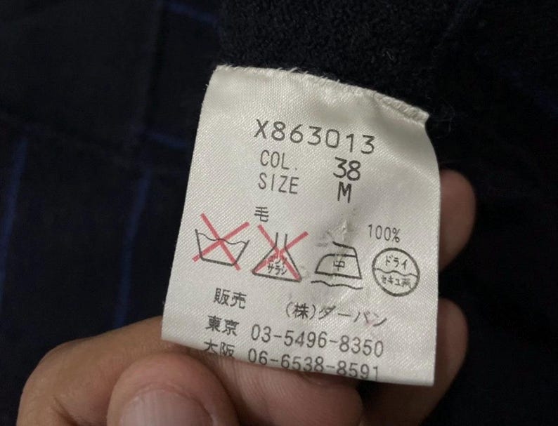 Puede incluir: Primer plano de una etiqueta de ropa con el texto "X863013", "COL. 38" y "SIZE M". La etiqueta tambi&eacute;n incluye s&iacute;mbolos de cuidado y texto japon&eacute;s. La tela es 100% lana.