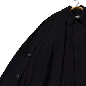 May include: A black cape-style coat with a collar and button closure. The coat is hanging on a wooden hanger. The sleeves have button details. The fabric appears to be a smooth, solid material.